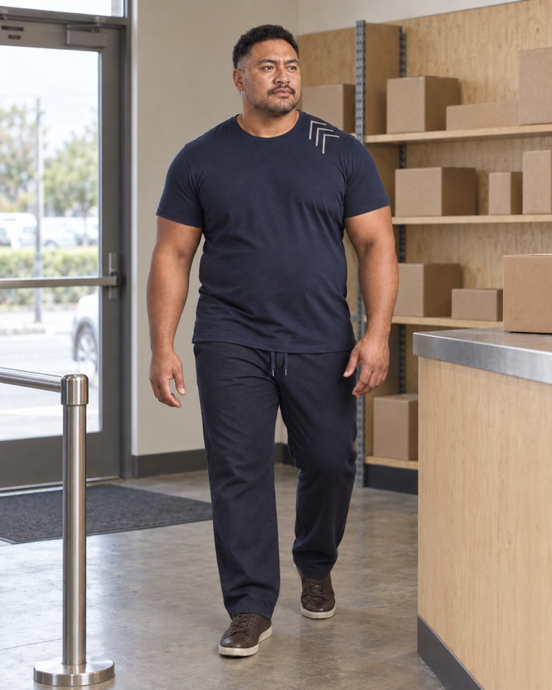 Deep Navy Oat Corner Gauge outfit worn at an ordinary neighborhood parcel pickup counter with plywood shelving, metal queue rail, cardboard boxes and a standard doorway
