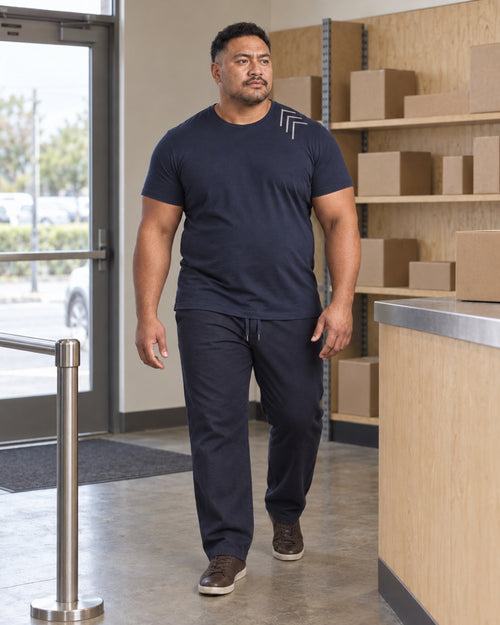Deep Navy Oat Corner Gauge outfit worn at an ordinary neighborhood parcel pickup counter with plywood shelving, metal queue rail, cardboard boxes and a standard doorway
